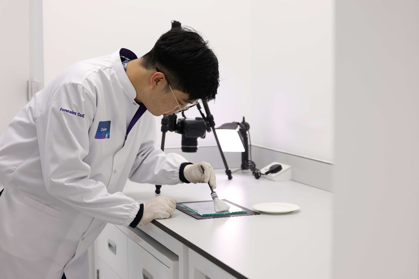 A man in a lab coat hunched over a desk, using a brush to dust a glass panel.